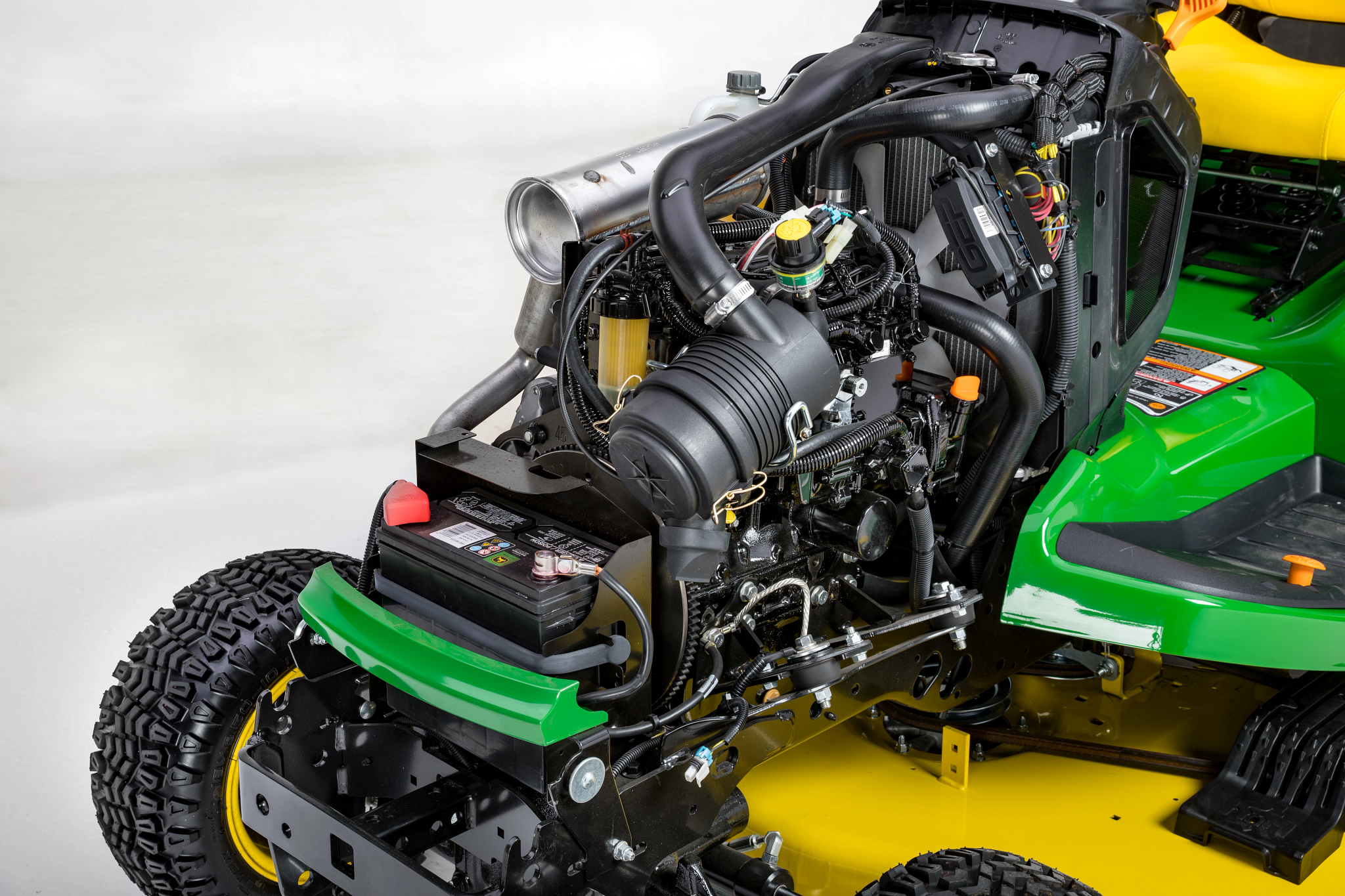 Close up of 3-Cylinder Diesel Engine lawn mower engine revealing battery, air intake, hoses, wiring, and other engine components mounted above a large treaded rear tire