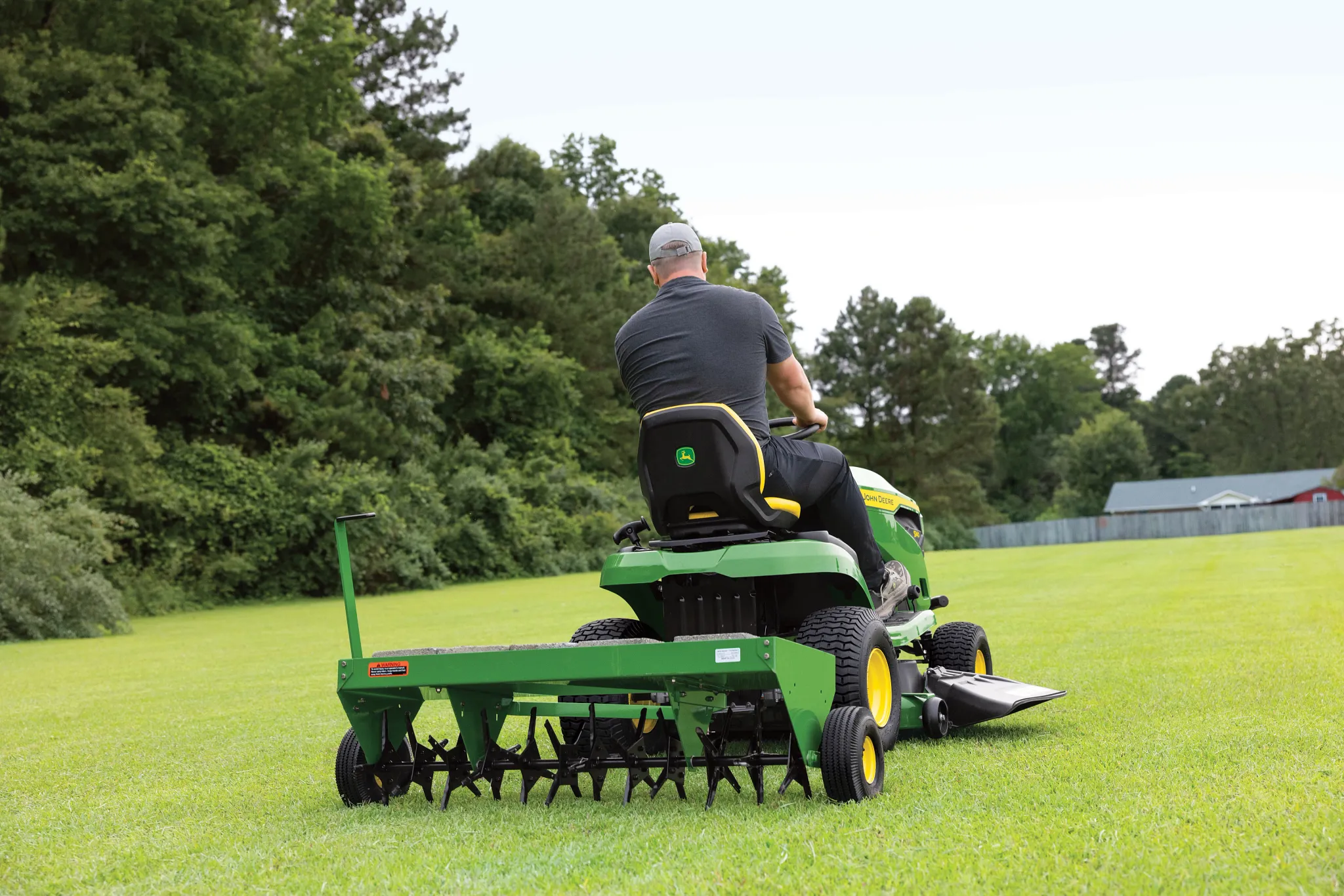 Rear view of an operator driving an S140 Lawn Tractor across a tree-lined lawn with attached aerator
