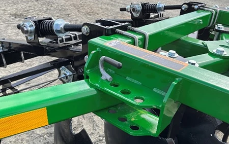 Close-up of agricultural implement frame showing a reinforced mounting bracket with a locking handle, bolt holes, warning label, and spring‑loaded adjustment components.