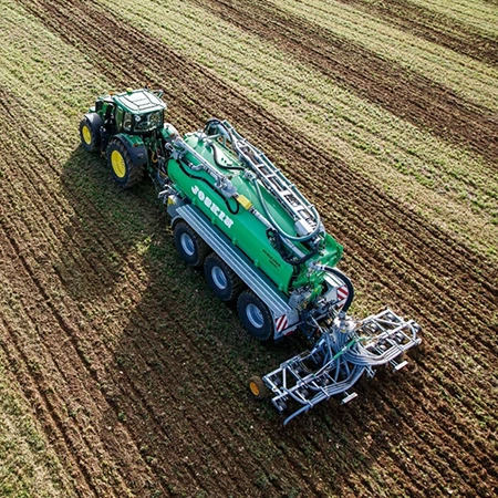 Overhead view of a green tractor pulling a large multi‑axle slurry tank and application implement across a field with parallel crop rows