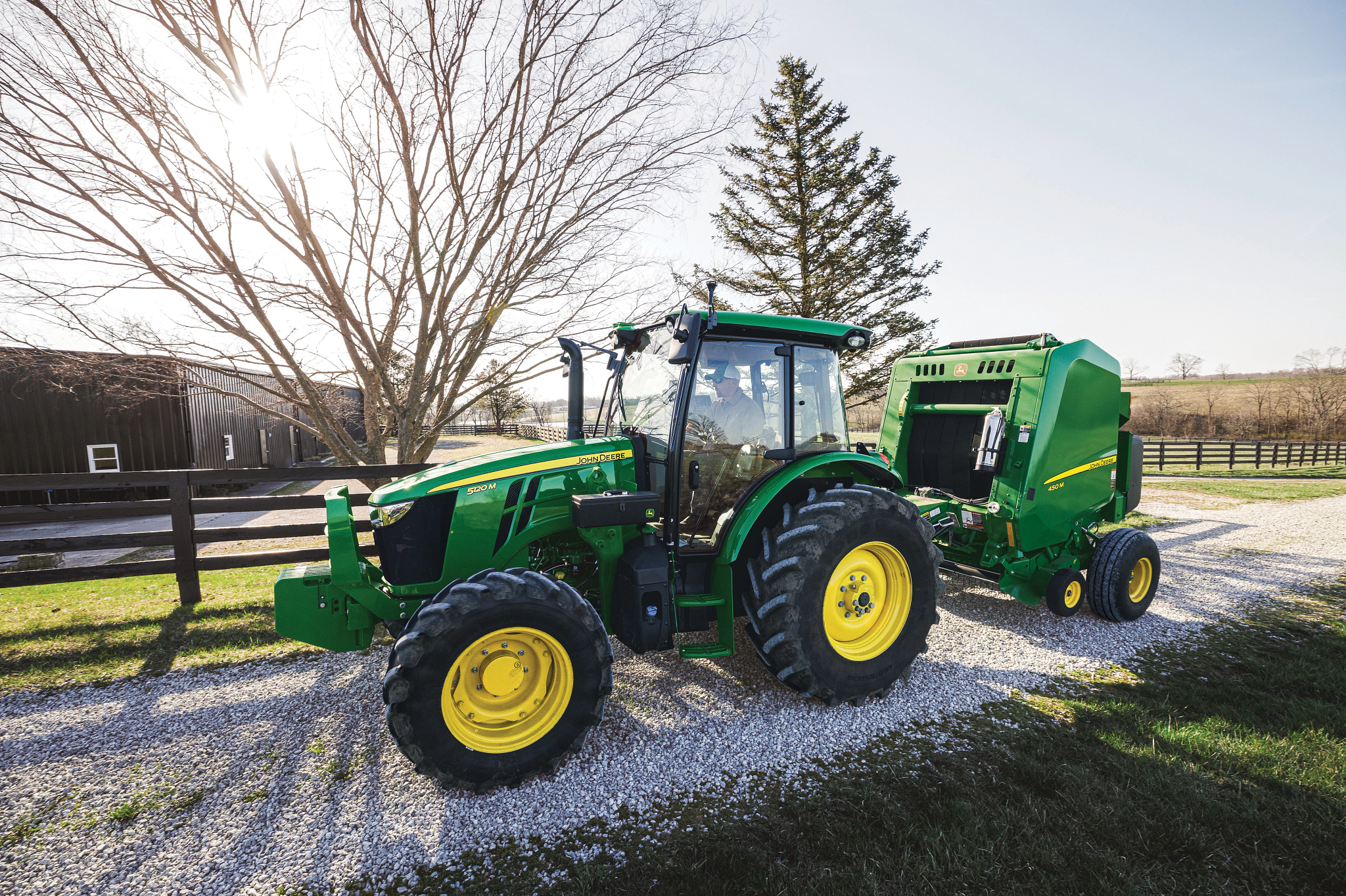 John Deere equipment consisting of a green tractor with enclosed cab pulling a green round baler on a gravel driveway near a farm fence, with trees and a barn in the background under late-day sunlight.