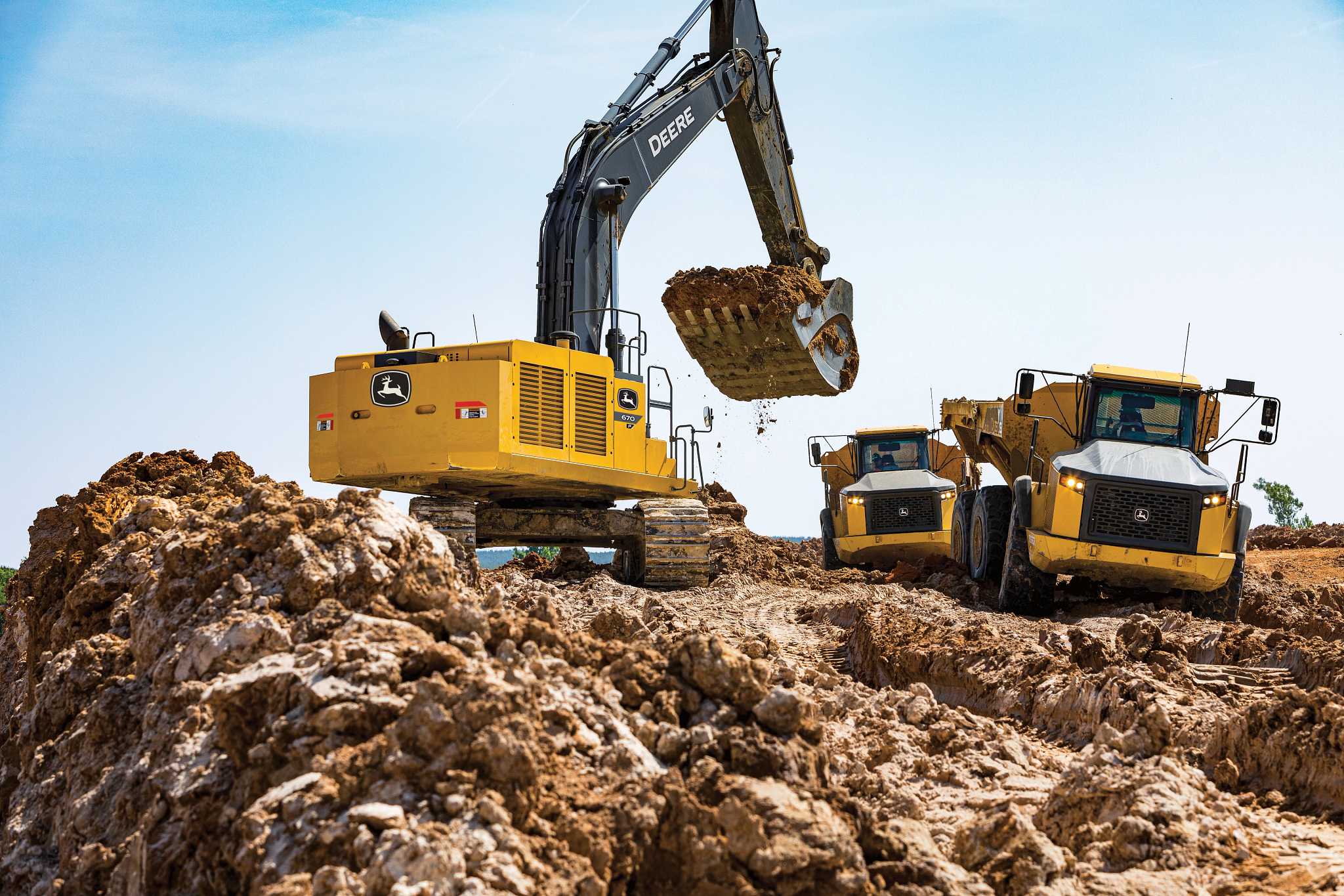 Yellow John Deere excavator with a black arm lifting a full bucket of dirt above a rough, muddy construction site