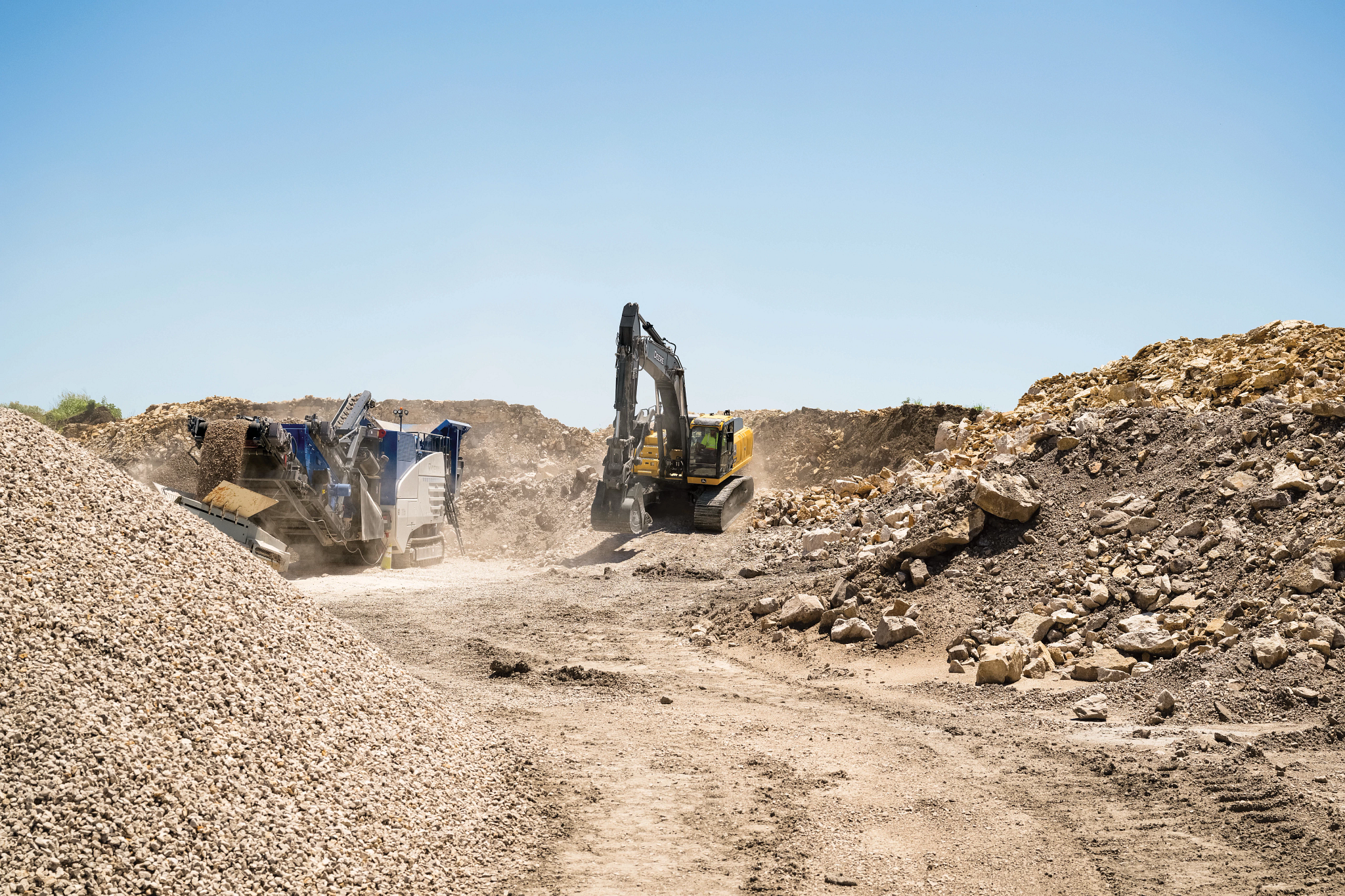 Wide view of a dusty quarry or construction site with piles of gravel and rocks, where a large yellow excavator and a rock-crushing machine operate along a dirt path, kicking up dust under a clear blue sky