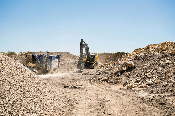 Wide view of a dusty quarry or construction site with piles of gravel and rocks, where a large yellow excavator and a rock-crushing machine operate along a dirt path, kicking up dust under a clear blue sky