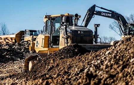 Large machine pushing a mound of dirt among rough soil piles and bare trees.