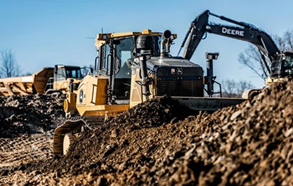 Large machine pushing a mound of dirt among rough soil piles and bare trees.