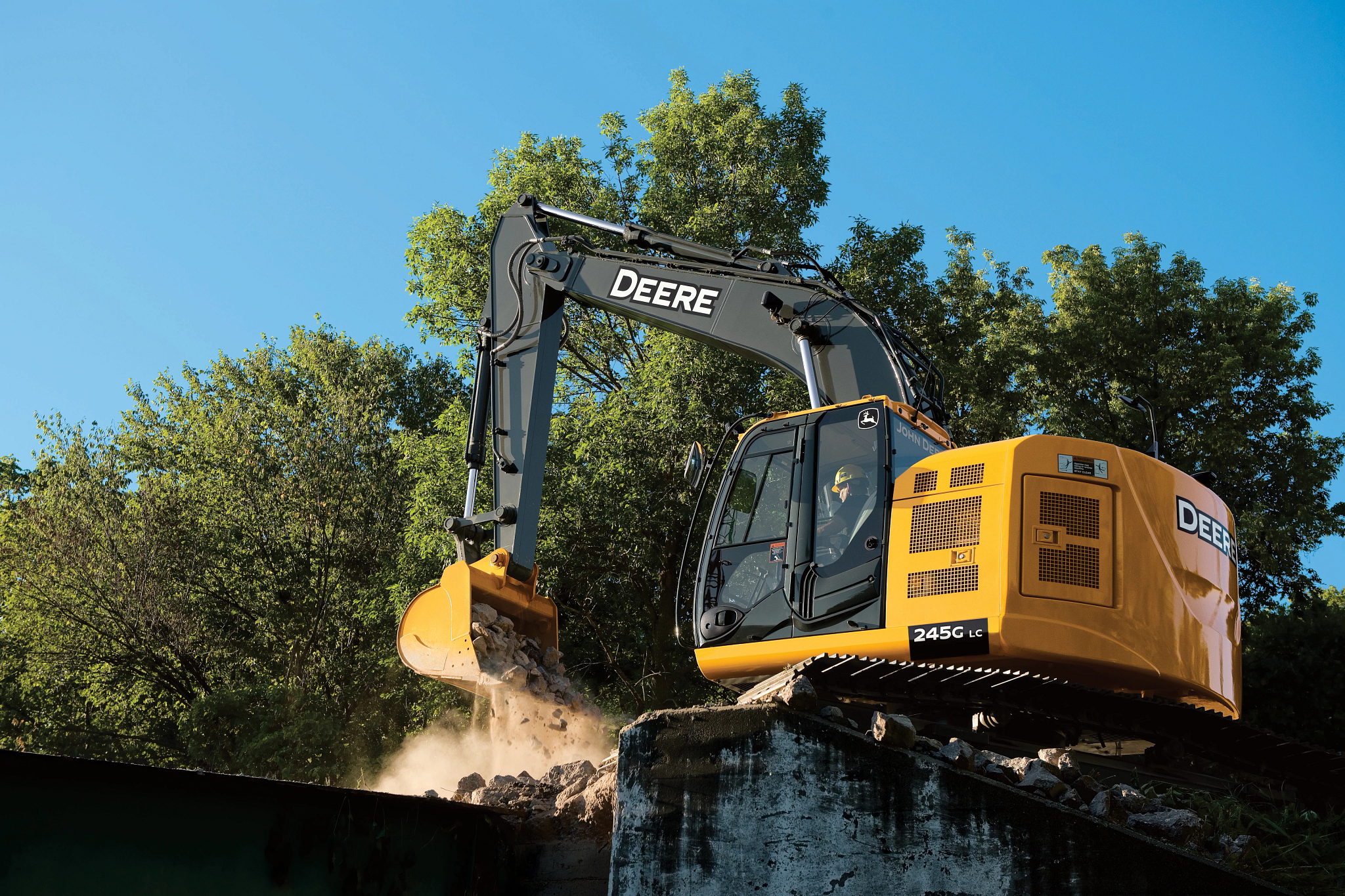 A yellow John Deere excavator sits on a raised concrete ledge, using its bucket to dump a load of rocks and debris over the edge