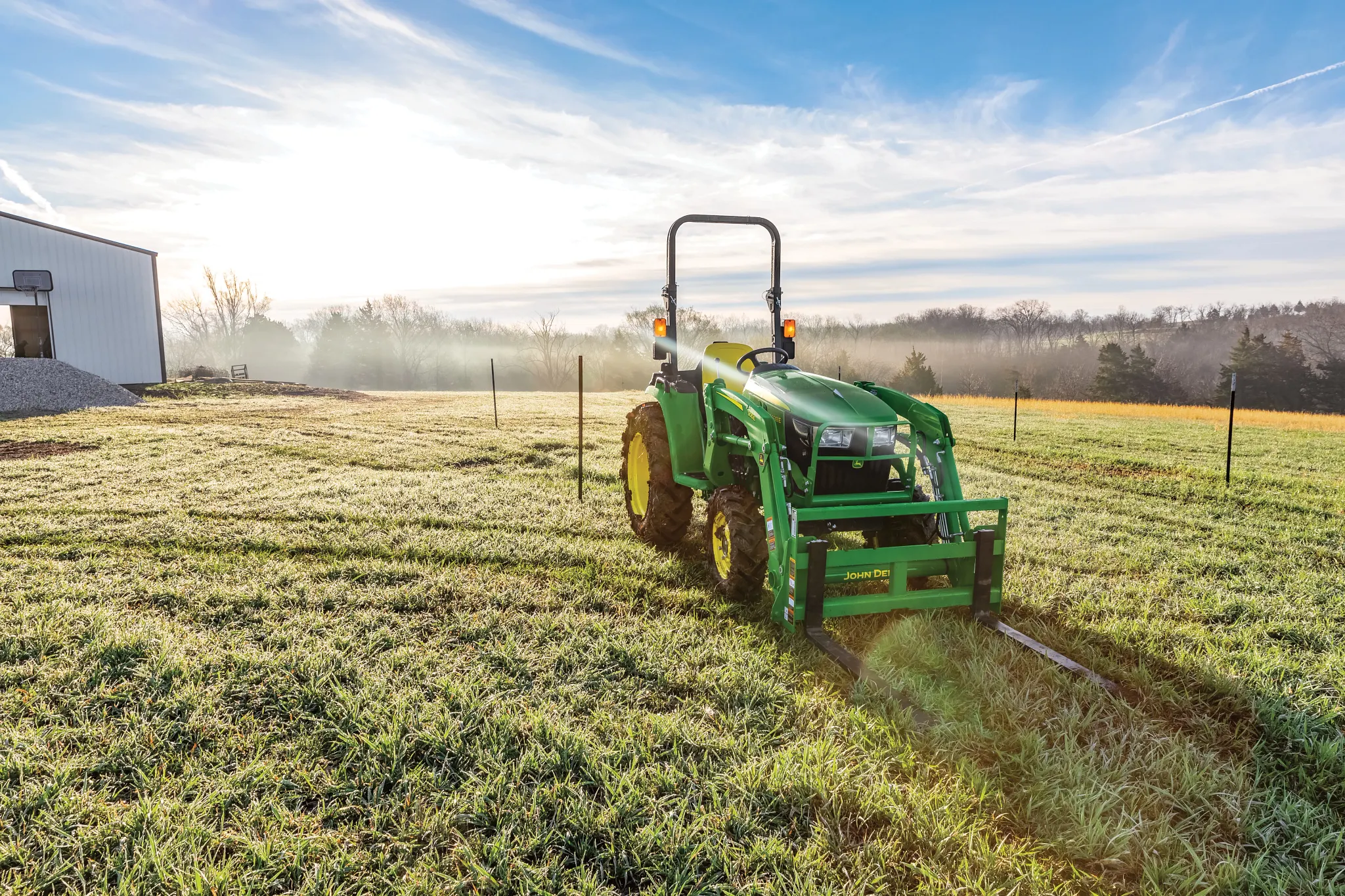 Green tractor parked in a frosty grass field as a building sits to the left under a bright early morning sky, with long shadows stretching across the ground and rolling landscapes in the background