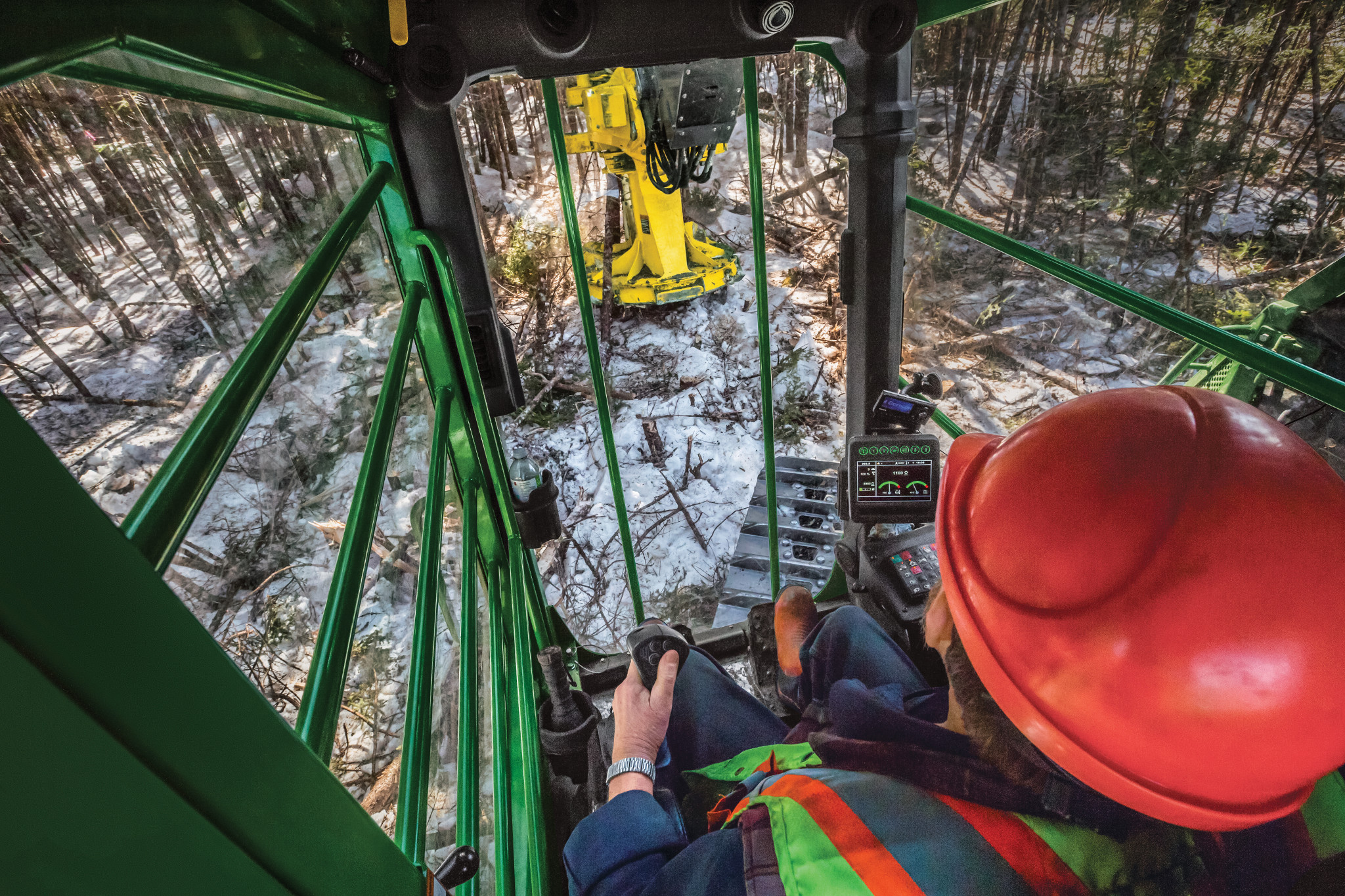 Inside cab view of man in florescent vest and safety helmet operating a tracked feller buncher in a snowy forest