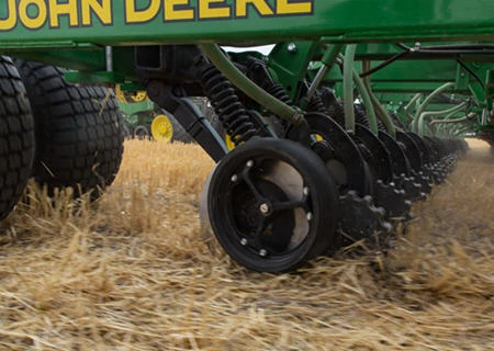Close-up of a John Deere tillage implement operating in a field, showing ground-engaging discs and gauge wheels moving through crop residue