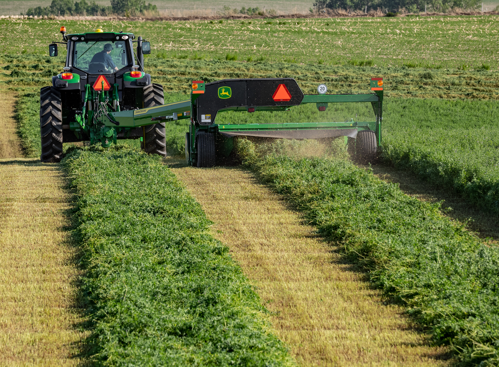 Rear view of a 6-series tractor with attached C400 mower-conditioner moving along rows of green crop