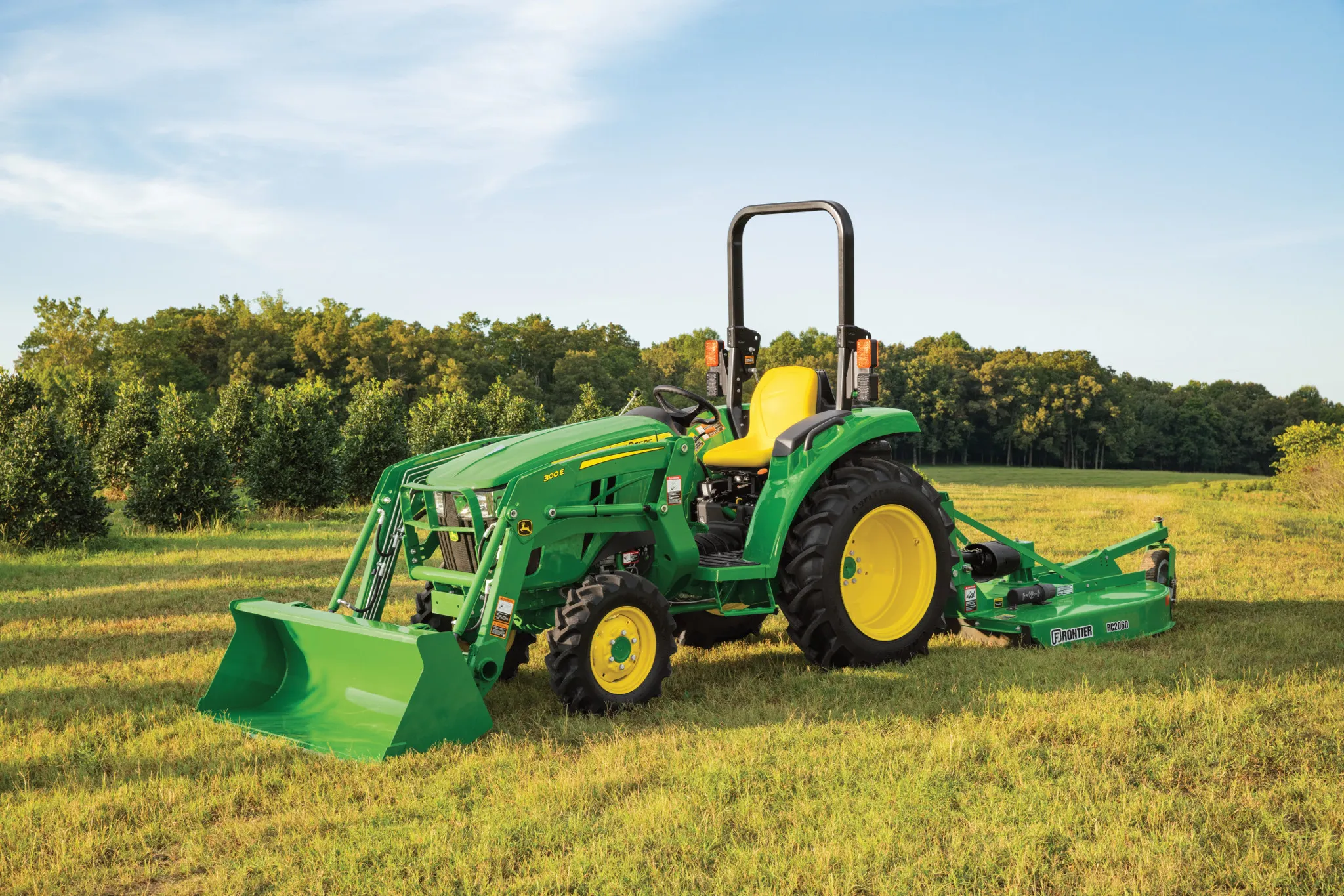A green tractor with a front loader and rear rotary cutter sits in a grassy field under a clear sky, with trees lining the background and sun shining down