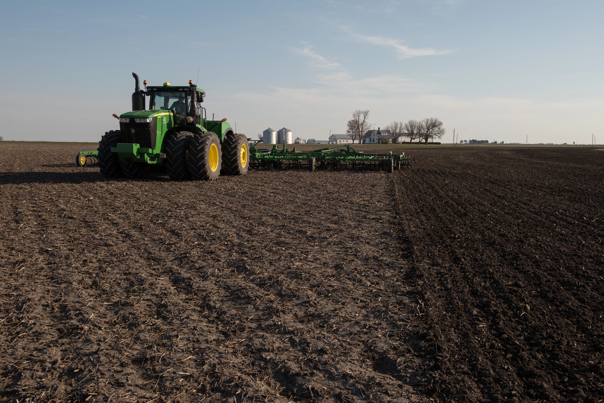 John Deere tractor pulling a field cultivator across a partially tilled field, with worked soil on the right, untouched residue on the left and farm buildings in the distance