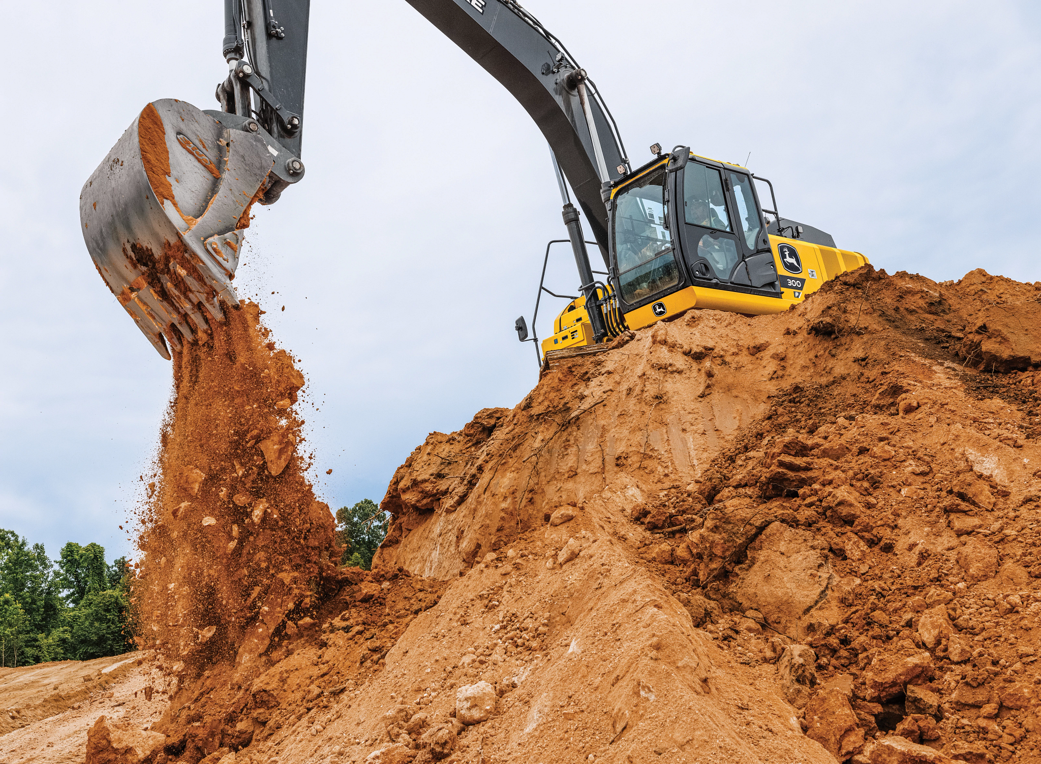 Yellow excavator on top of a sandy dirt mound using its bucket to dump a cascade of loose soil down the side of the pile at a construction site under a cloudy sky.