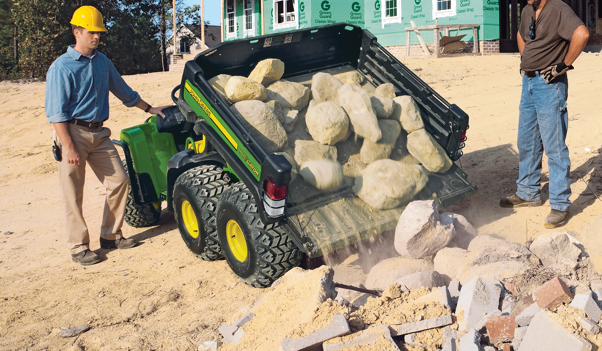 Rear view of a TH 6x4 Gas Gator dumping boulders from its cargo box while two construction workers look on