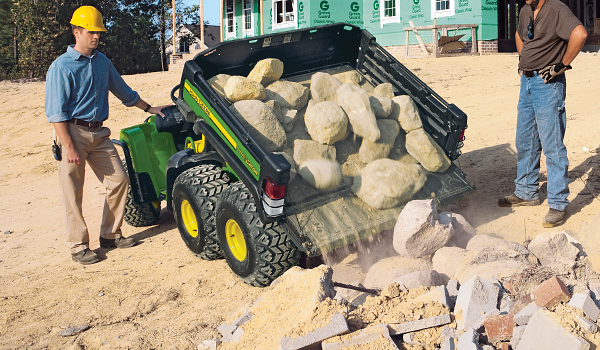 Rear view of a TH 6x4 Gas Gator dumping boulders from its cargo box while two construction workers look on