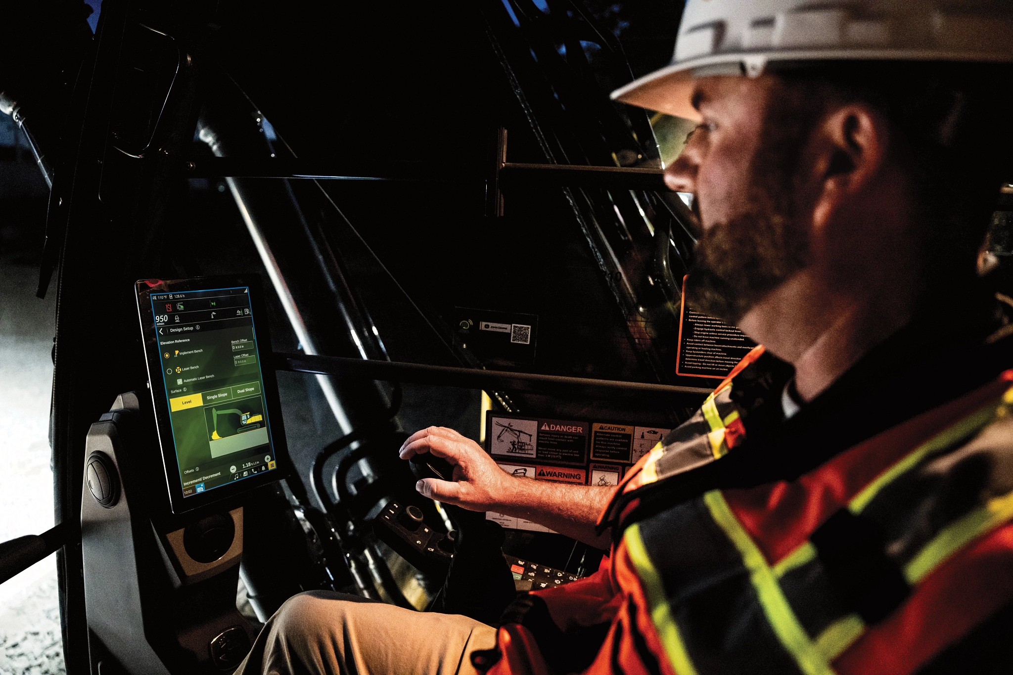 A construction worker wearing a hard hat and high-visibility vest operates heavy machinery from an enclosed cab at night