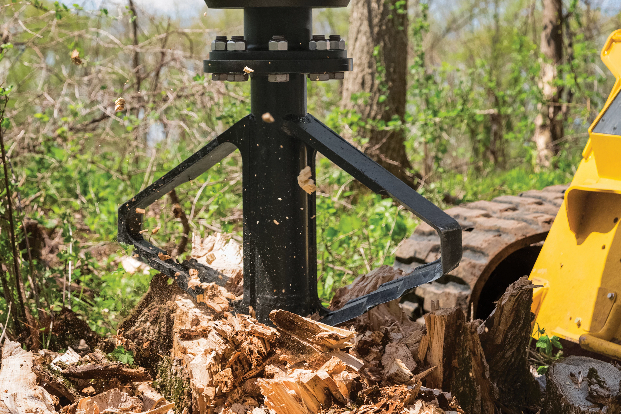 Close-up of a stump shredder attachment drilling into a tree stump, with rotating metal blades breaking wood and debris in a wooded setting