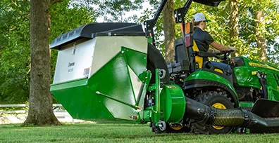 MAn operating a green mower equipped with a rear-mounted lawn debris collection unit and vacuum hose in a park-like setting