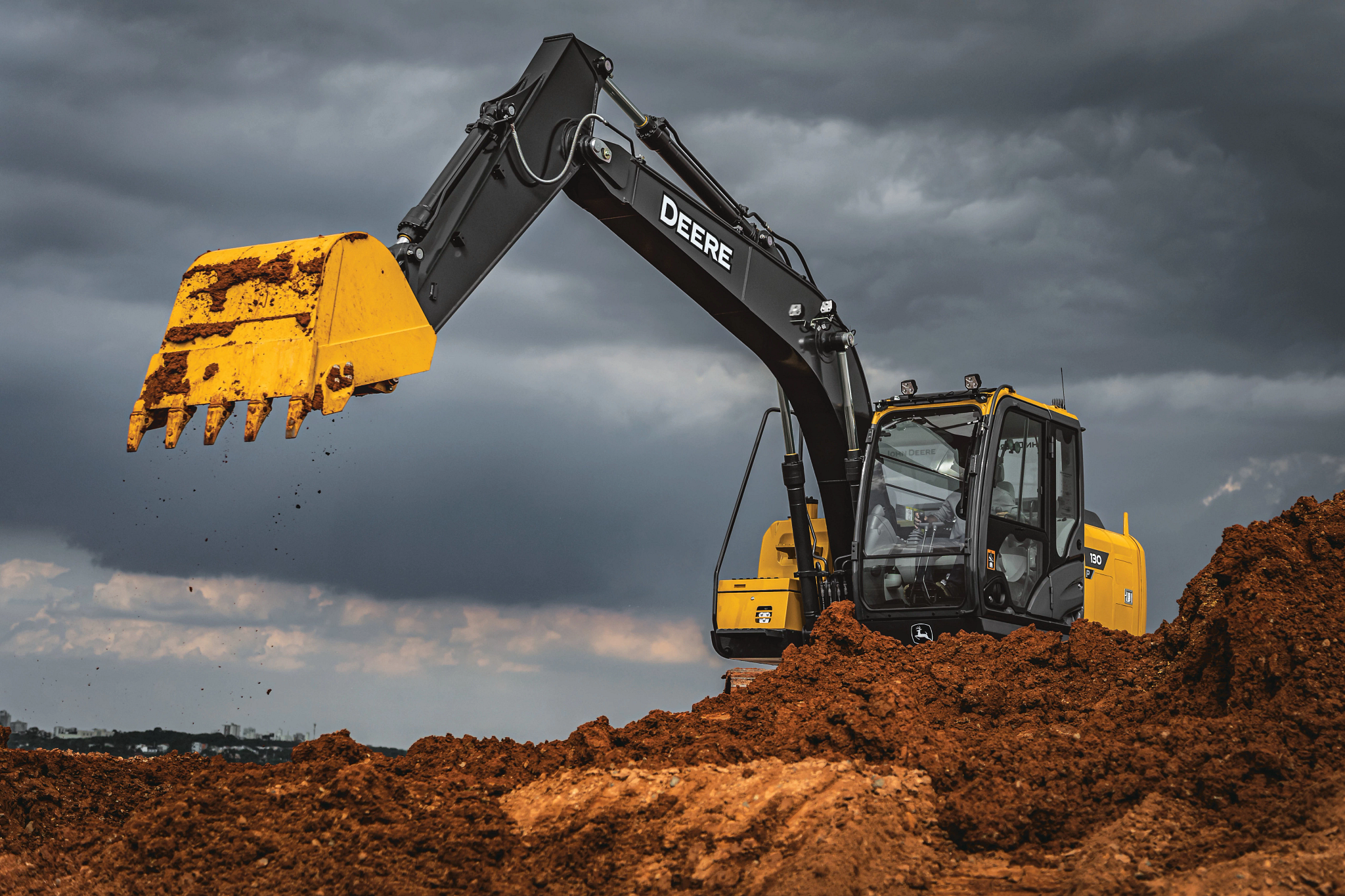 Yellow and black excavator positioned on a mound of reddish-brown soil, lifting a large bucket as small clumps of dirt fall from the teeth, with dark, stormy clouds filling the sky in the background
