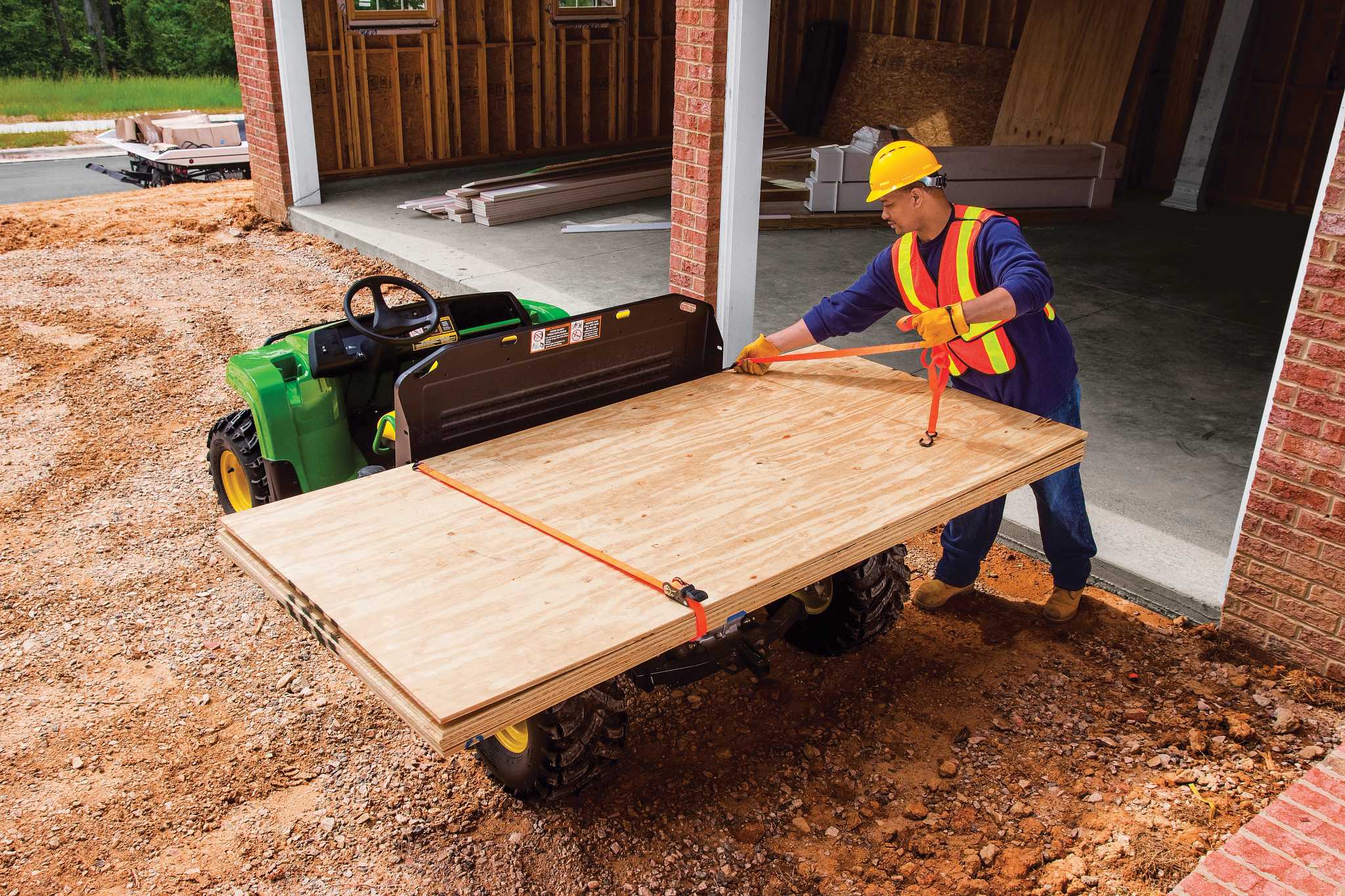 Hard hat-wearing worker straps a large sheet of plywood to the top of the cargo box of a TX 4X2 Gator in front of a new garage build