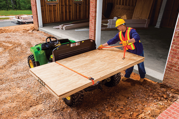 Hard hat-wearing worker straps a large sheet of plywood to the top of the cargo box of a TX 4X2 Gator in front of a new garage build