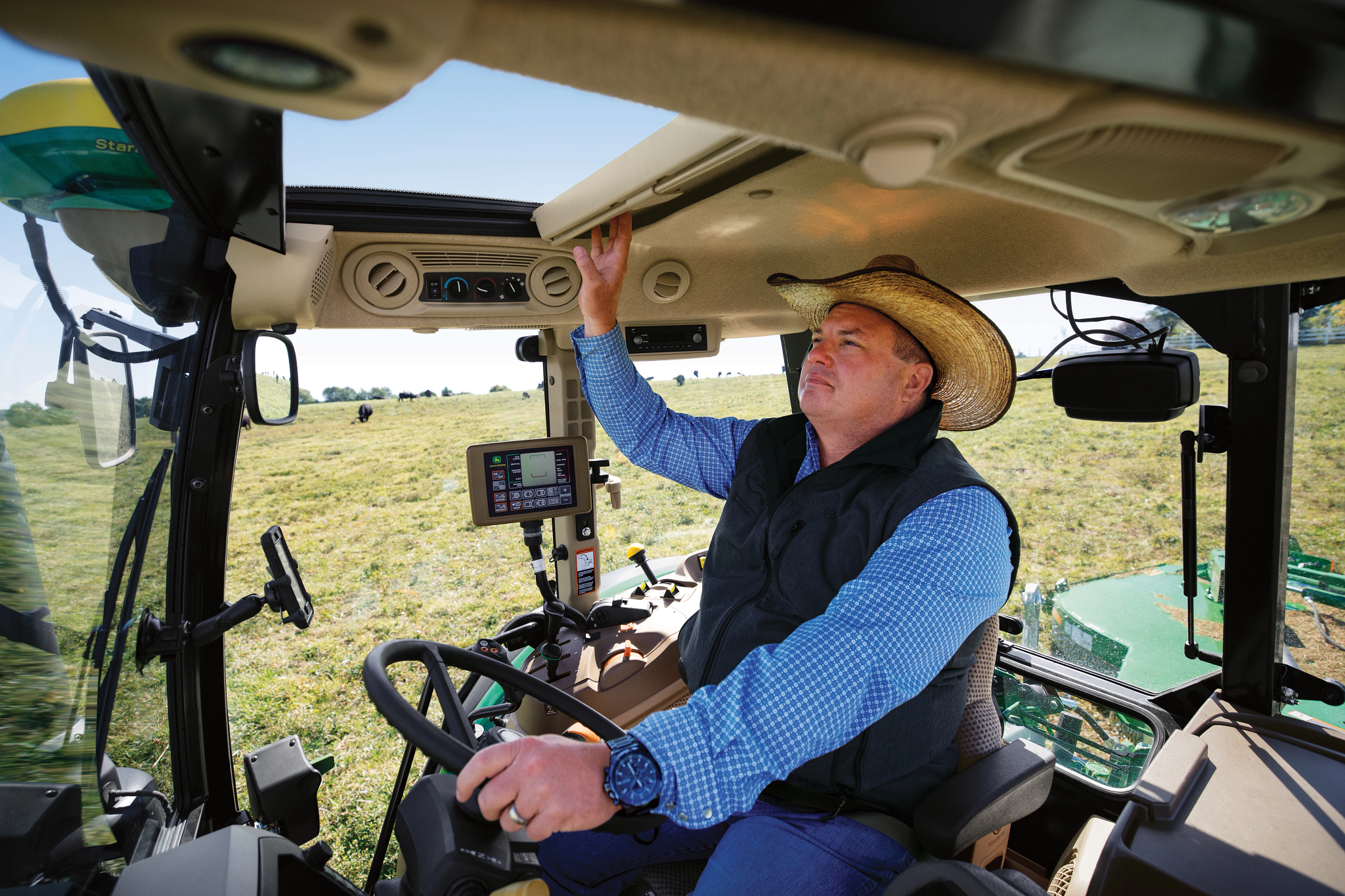 Operator seated inside a tractor cab, holding the steering wheel with one hand and adjusting an overhead control with the other