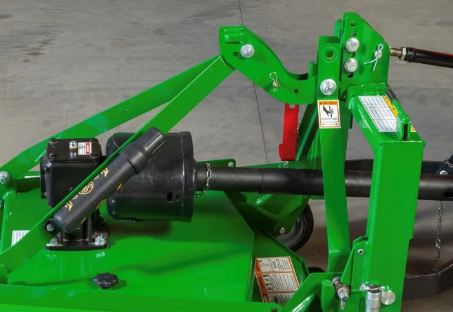 Close‑up of a green grooming mower showing a PTO driveline, gearbox, and metal linkage components