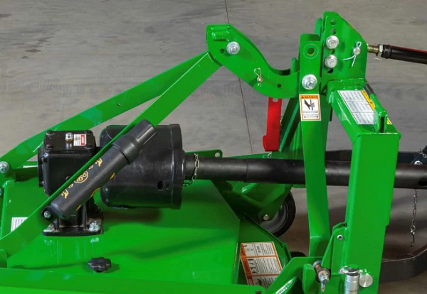 Close‑up of a green grooming mower showing a PTO driveline, gearbox, and metal linkage components