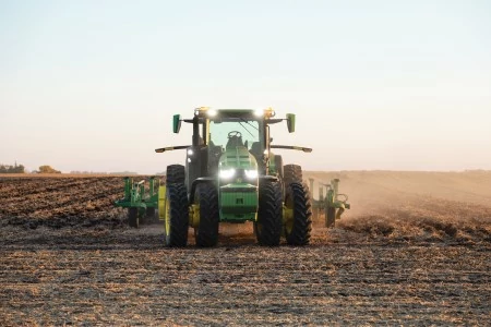 Front view of a green tractor driving across a harvested field at sunset, with large rear tires, illuminated headlights, and soil dust rising behind attached tillage equipment.