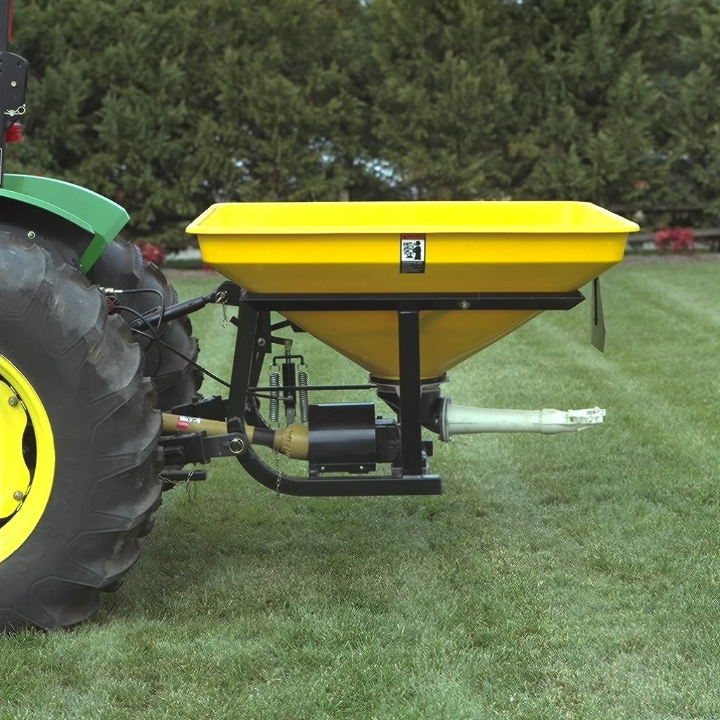Side view of a yellow broadcast spreader mounted on the rear of a tractor, positioned on a grassy lawn with trees in the background