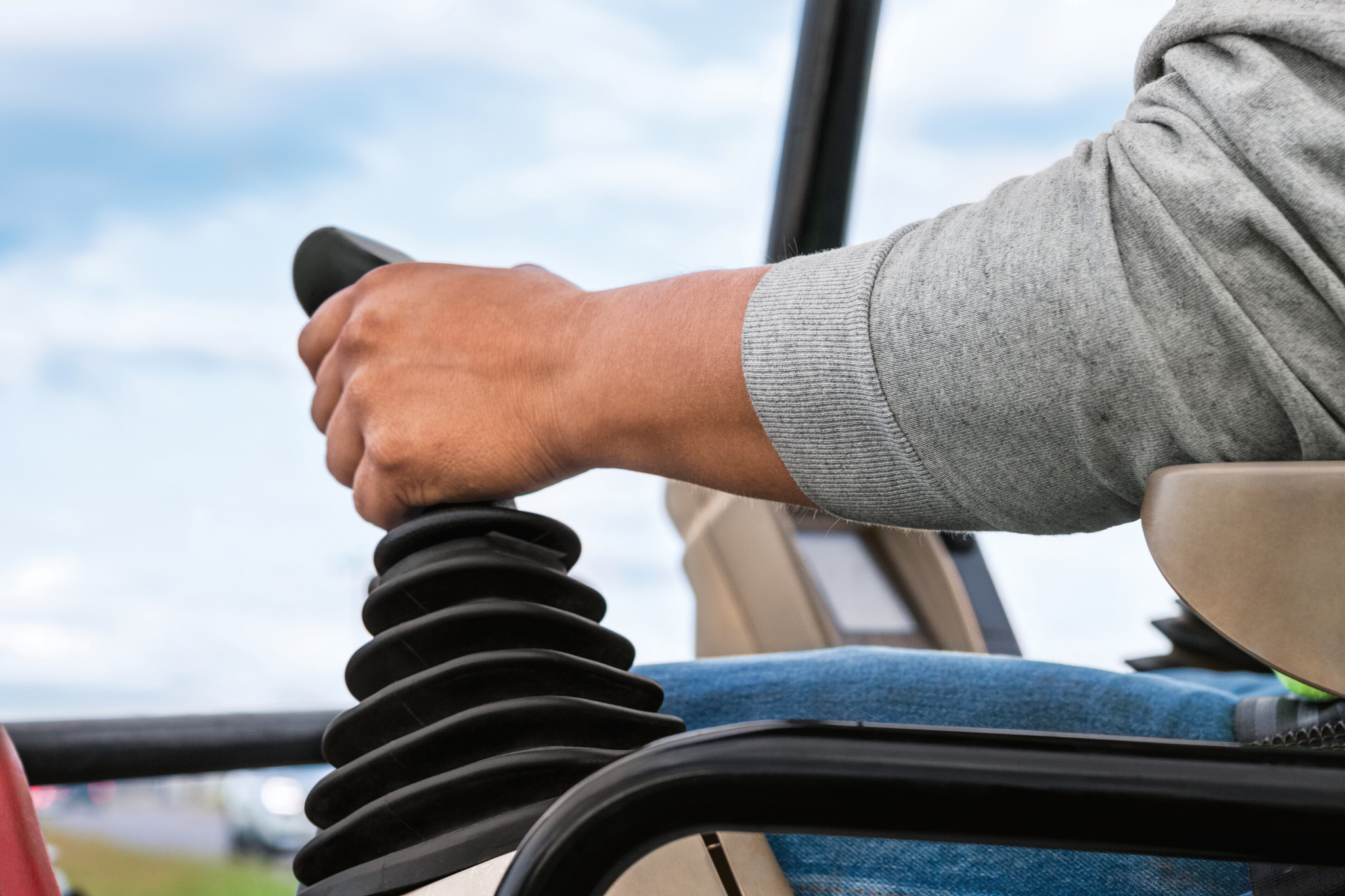 Close-up of a person’s hand gripping a joystick control lever inside heavy equipment, with a cloudy sky visible through the window in the background