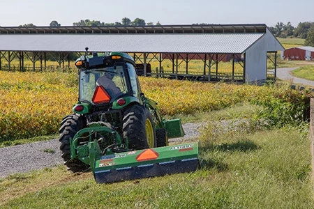 Rear view of a cab tractor using a flail mower attachment on a grassy hill in front of a yellow field and long, side-less farm building