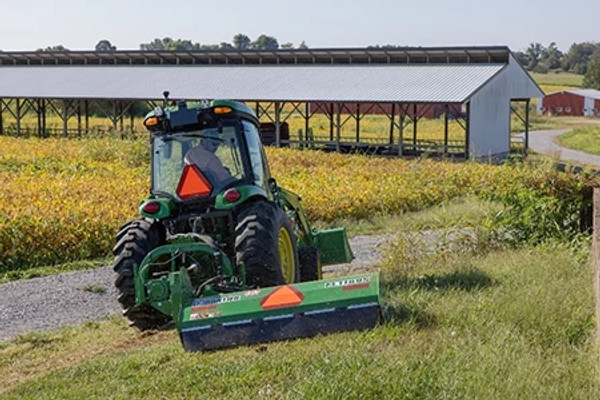 Rear view of a cab tractor using a flail mower attachment on a grassy hill in front of a yellow field and long, side-less farm building