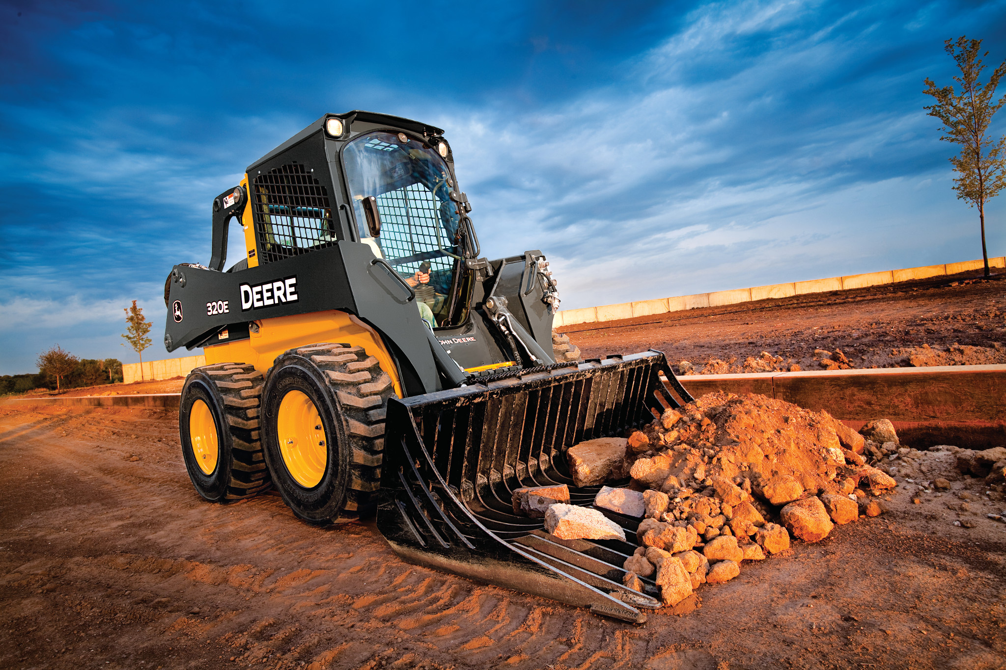 A compact skid-steer loader with “DEERE” branding uses a rock bucket attachment to scoop a pile of large stones and dirt on a construction site, with tire tracks visible in the soil, a low retaining wall in the background, and a clear blue sky above.