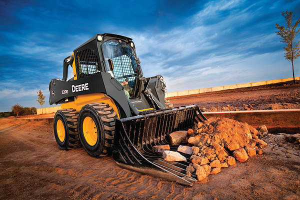 A compact skid-steer loader with “DEERE” branding uses a rock bucket attachment to scoop a pile of large stones and dirt on a construction site, with tire tracks visible in the soil, a low retaining wall in the background, and a clear blue sky above.