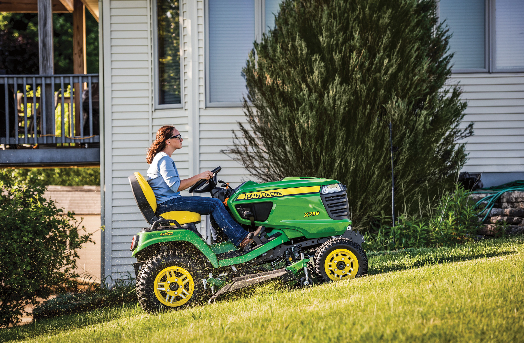 Side right view of a homeowner driving an X739 Signature Series Tractor with 60-in. mower deck up a hilly lawn with a house in the background