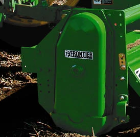 Close‑up of a green Frontier agricultural mower attachment showing the curved protective side panel with the Frontier logo, mounted low to the ground with crop residue and soil around the cutting area.