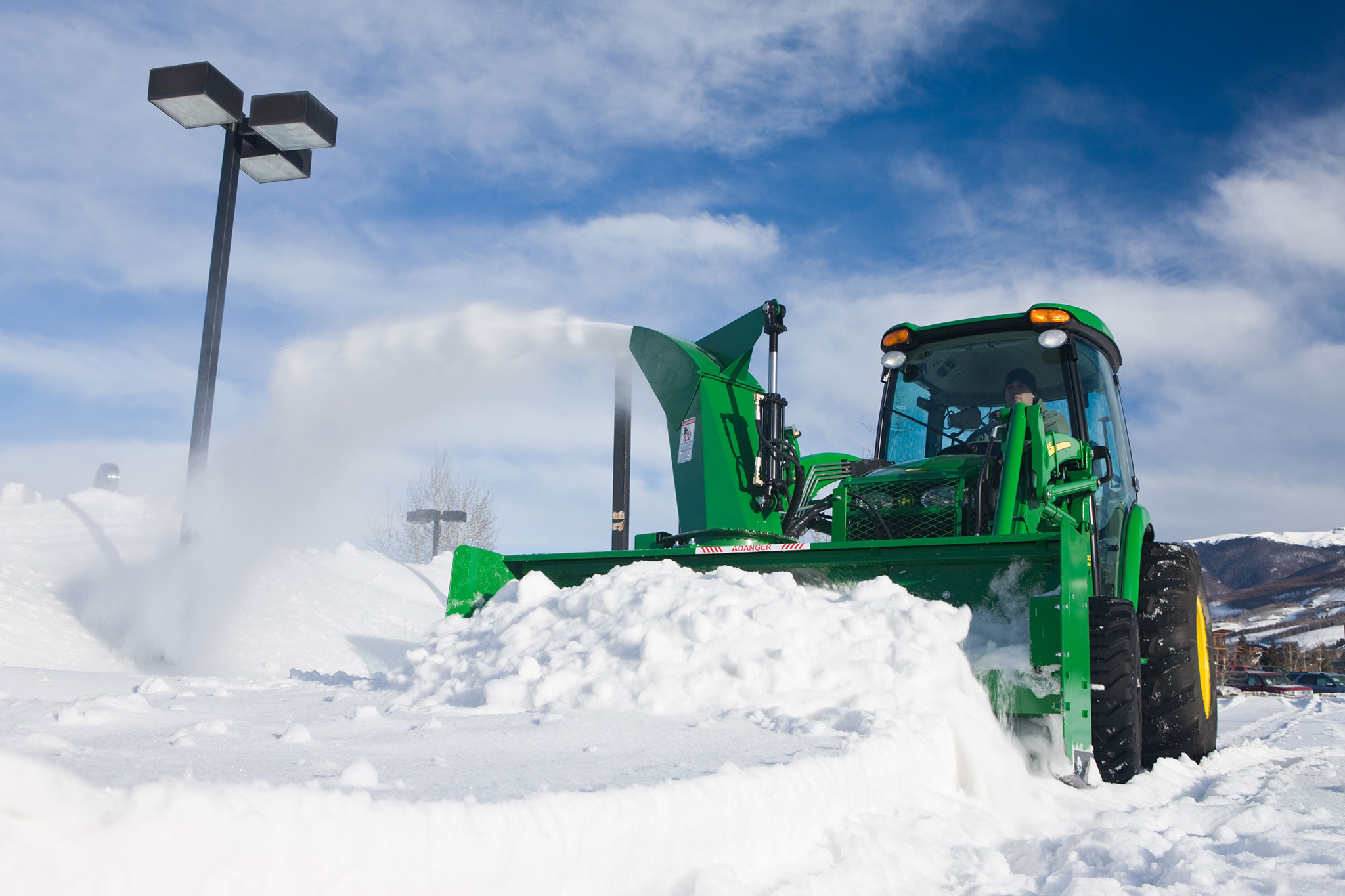 Tractor equipped with a front-mounted snow blower clearing a large pile of snow, with snow blowing out of the chute against a blue sky