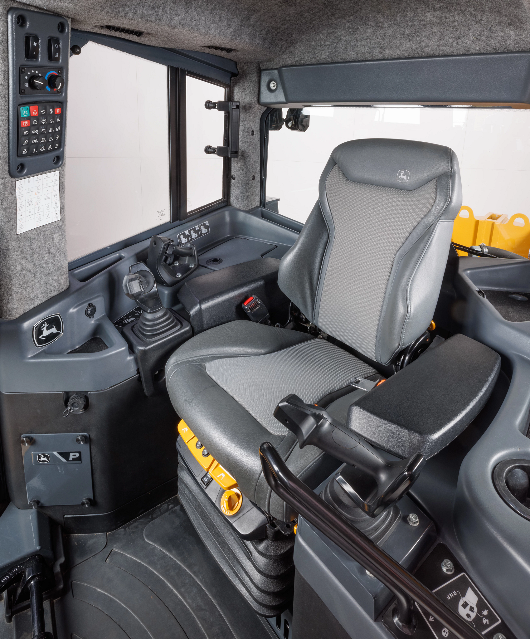 Interior of equipment or tractor cab showing a gray cushioned operator seat, joystick controls, armrest, floor pedals, side control panels with buttons and climate dials, and large cab windows.