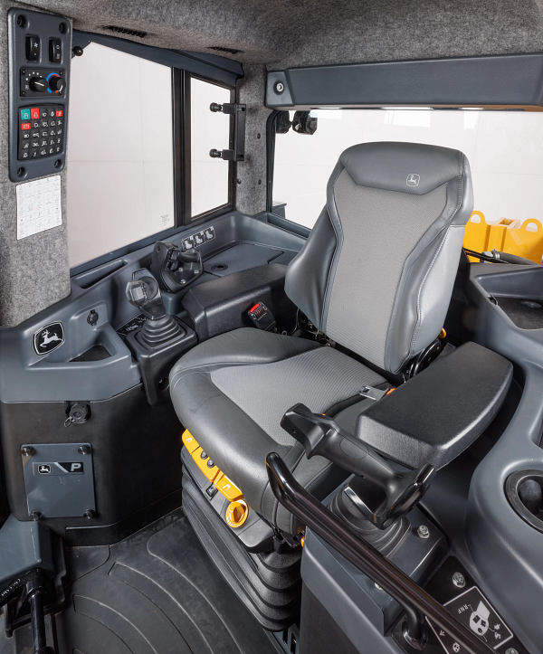 Interior of equipment or tractor cab showing a gray cushioned operator seat, joystick controls, armrest, floor pedals, side control panels with buttons and climate dials, and large cab windows.
