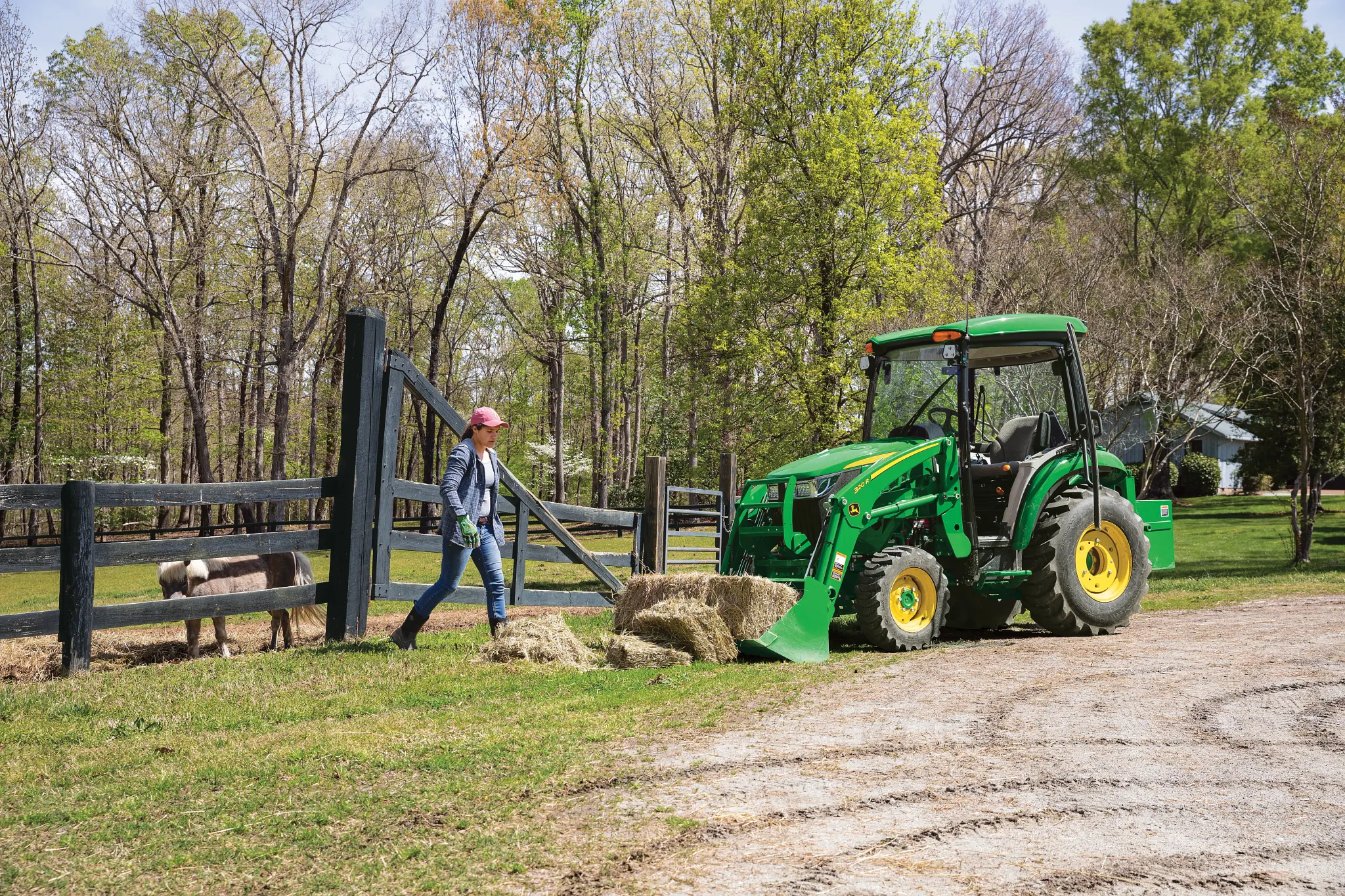 Green 320R tractor parked alongside a horse pen, with driver walking toward it to pick up hay bales in its front loader bucket