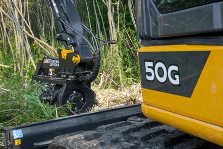 John Deere 50G compact excavator equipped with a hydraulic forestry mulching head, shown clearing vegetation along a wooded edge