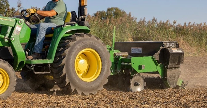 Tractor pulling a rear‑mounted tillage or soil‑preparation attachment across a field, with the implement churning dry soil and residue.