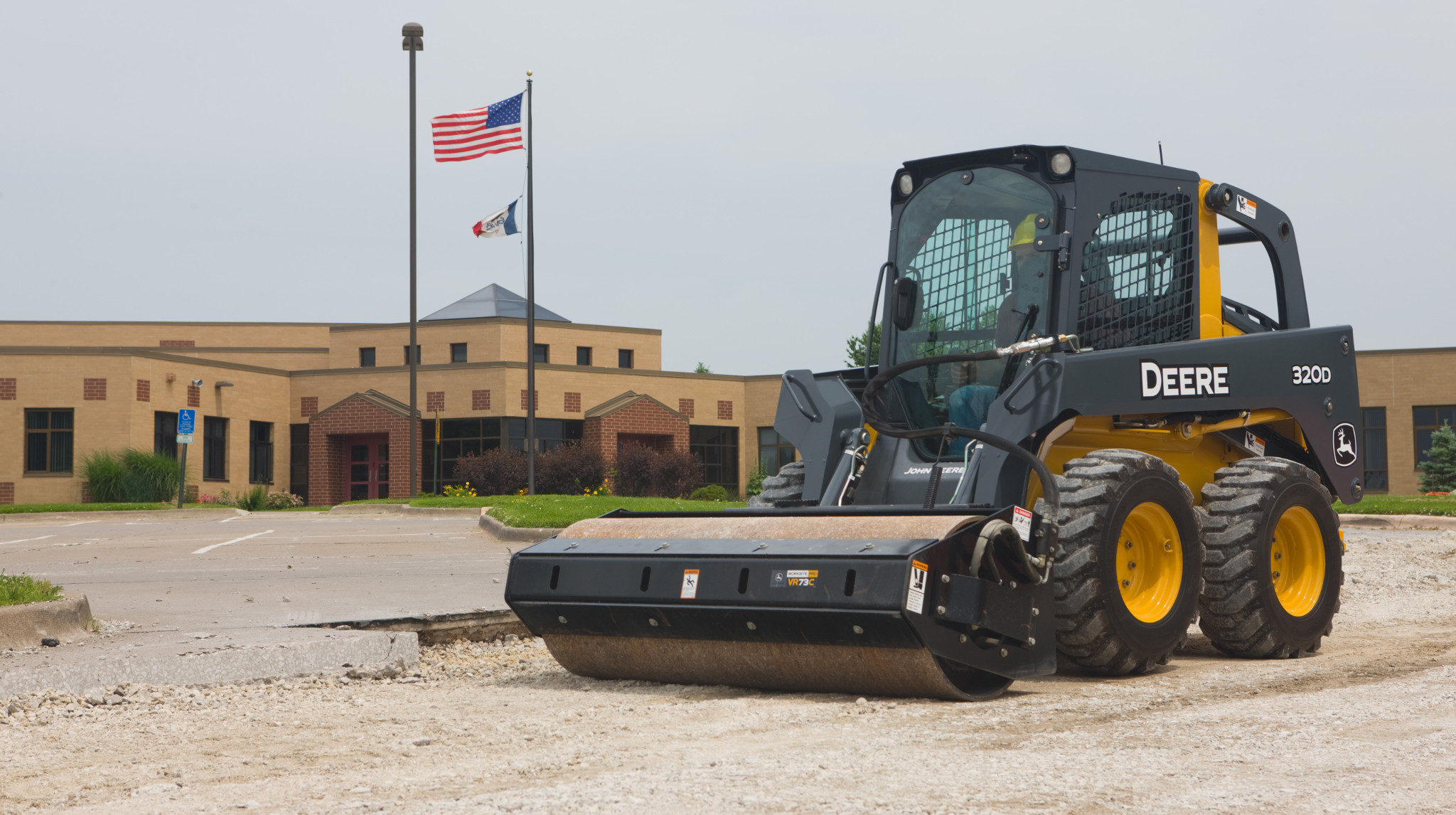 Front left view of VR Series Vibratory Roller on a 320D Skid Steer rolling a gravel section of parking lot outside a large building
