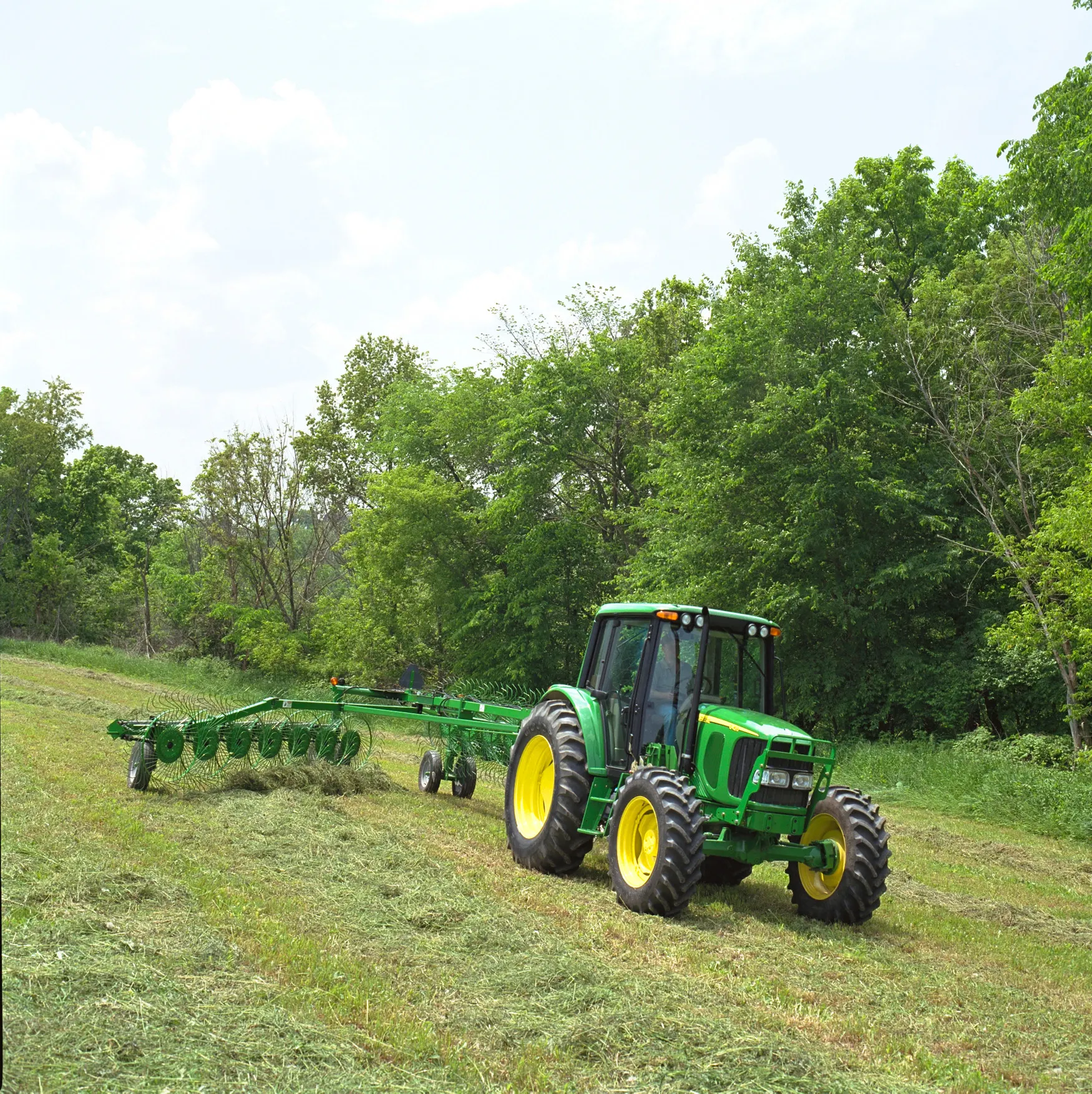 Front right, angled view of a tractor pulling a WR High-Capacity Wheel Rake across a hay field bordered by trees