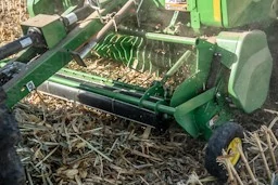 John Deere corn header operating over harvested corn residue, with rotating intake and cutting components processing stalks on the ground.