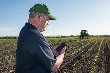 Man in a blue shirt and ballcap looking at a cell phone while a sprayer operates in the background