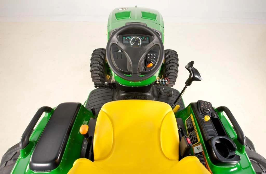 Operator’s view of a John Deere lawn tractor showing yellow seat, steering wheel with instrument cluster, hydrostatic control lever, cup holder, and large rear tires on a clean studio background.