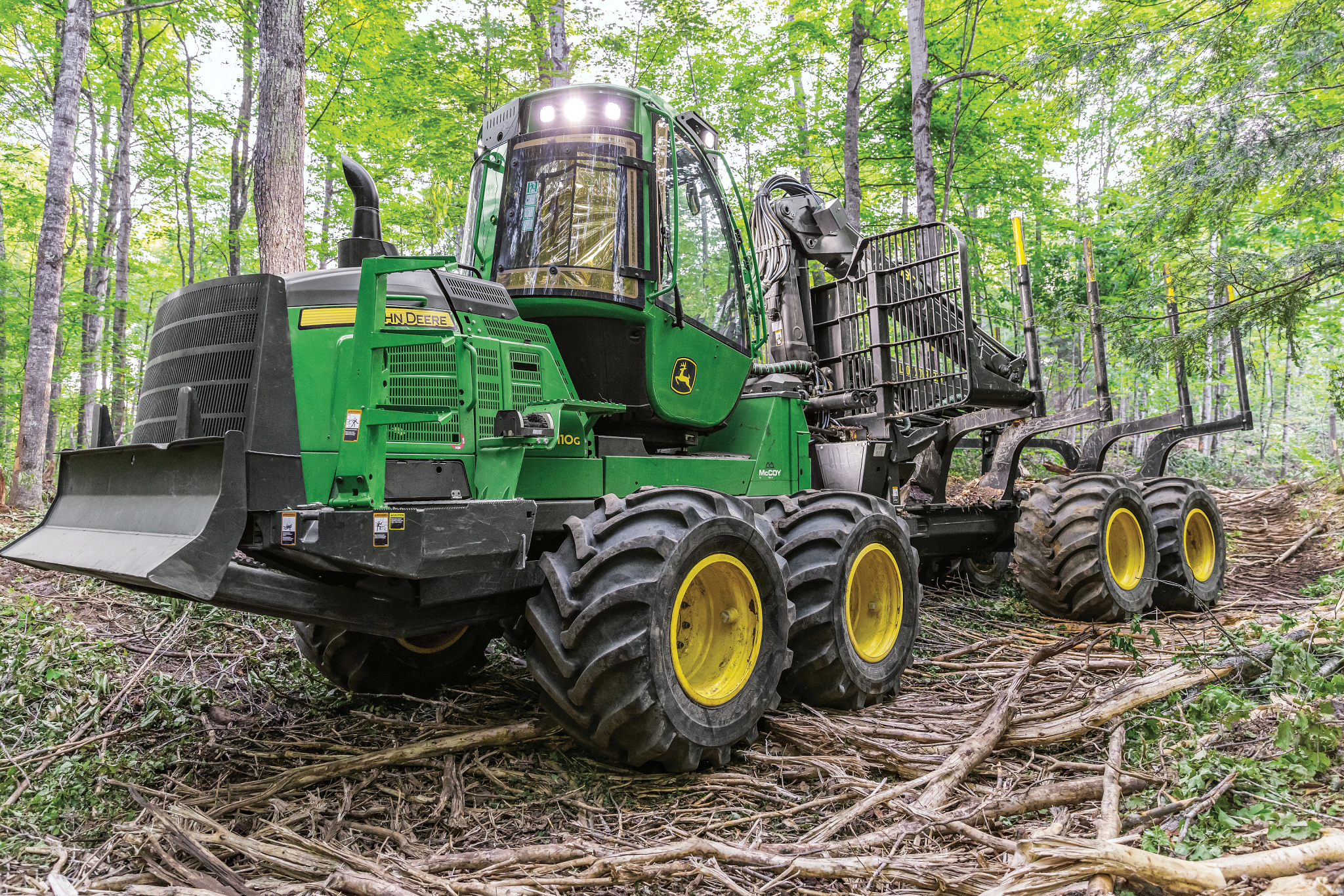 A green forestry forwarder with large articulated tires in a wooded area, carrying a rear log‑loading cradle and surrounded by fallen branches